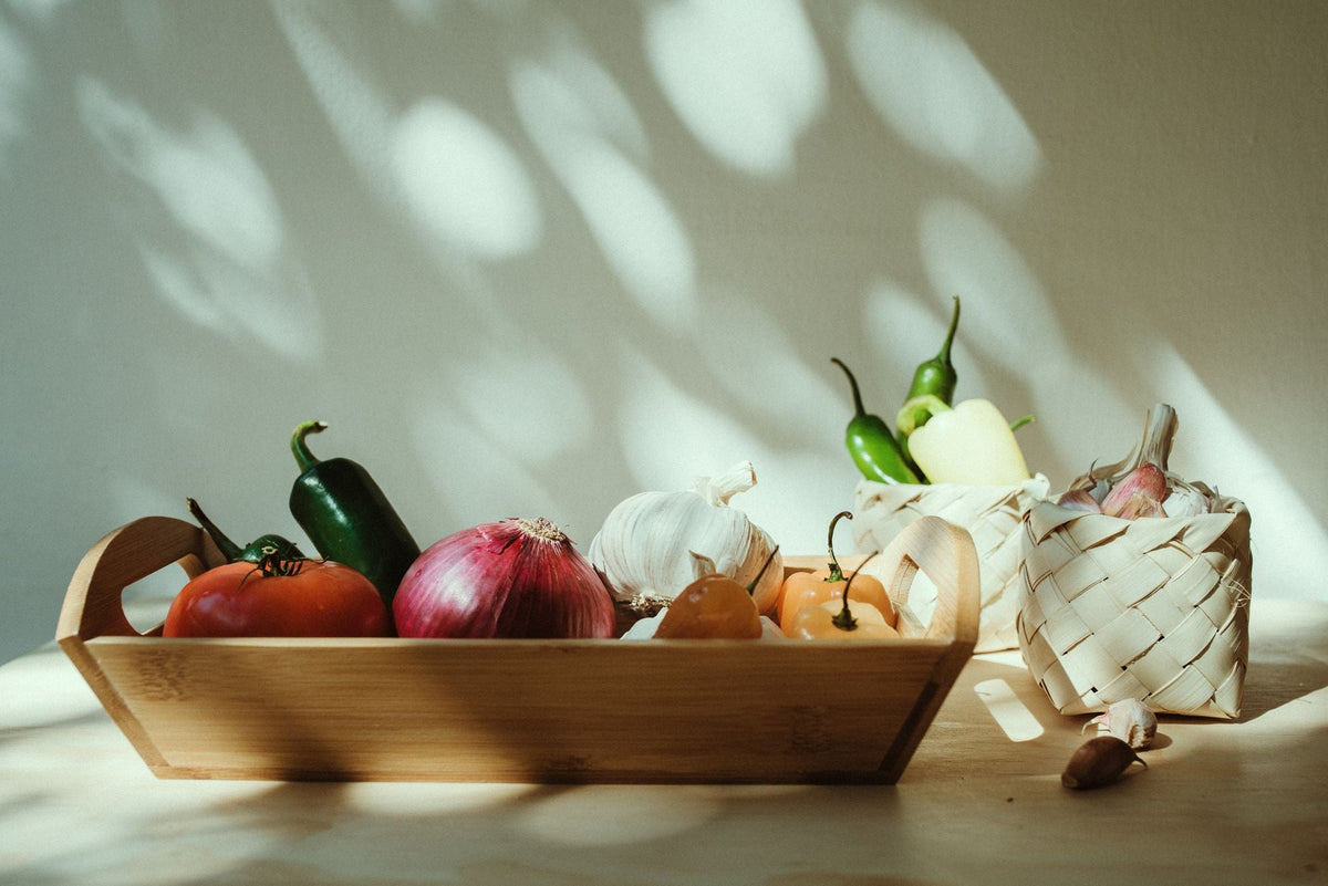 Charola de madera con verduras frescas bajo luz natural que proyecta sombras de hojas en la pared