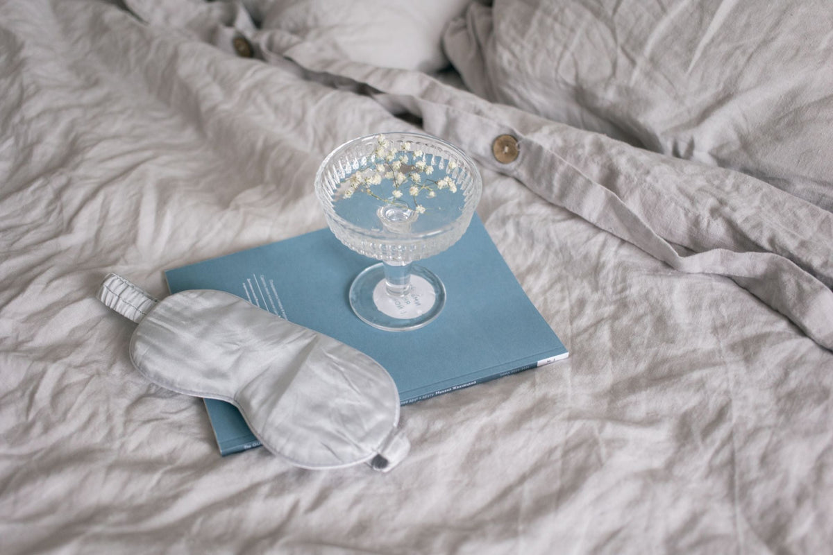 Antifaz para dormir, libro y copa con flores secas sobre sábanas de lino arrugadas