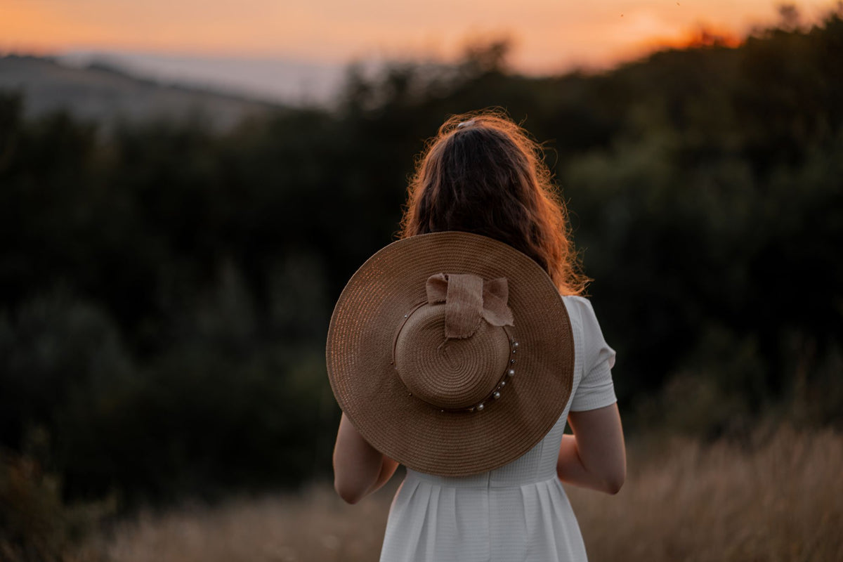 Mujer de espaldas con vestido blanco y sombrero de paja en la mano, en una tarde de verano en el campo